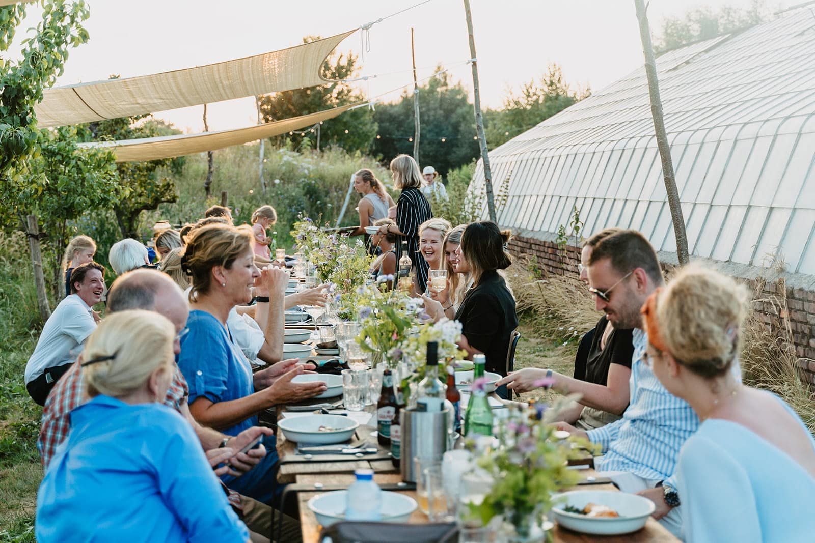 Gesellige Hochzeitsgesellschaft an einer langen Tafel im Grünen während der Abendstunden eines Hochzeitswochenendes