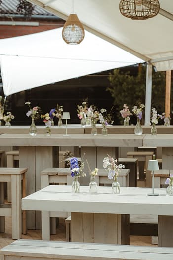 Minimalistisch und modern gedeckte Tafel mit feinen Glasvasen und Sommerblumen unter einer Zeltplane – Teil einer hochwertig geplanten Outdoor- und Zelthochzeit.