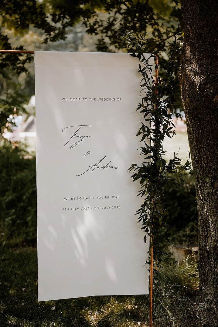 Minimalistisches Welcome-Schild für ein Wedding Weekend an einem Airbnb-Ferienhaus, platziert im Grünen bei einer stilvollen Outdoor-Hochzeit.