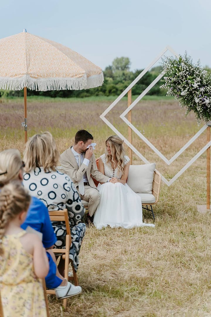 Brautpaar sitzt bei einer freien Trauung mitten in der weiten unbebauten Natur, umgeben von Familie und Freunden