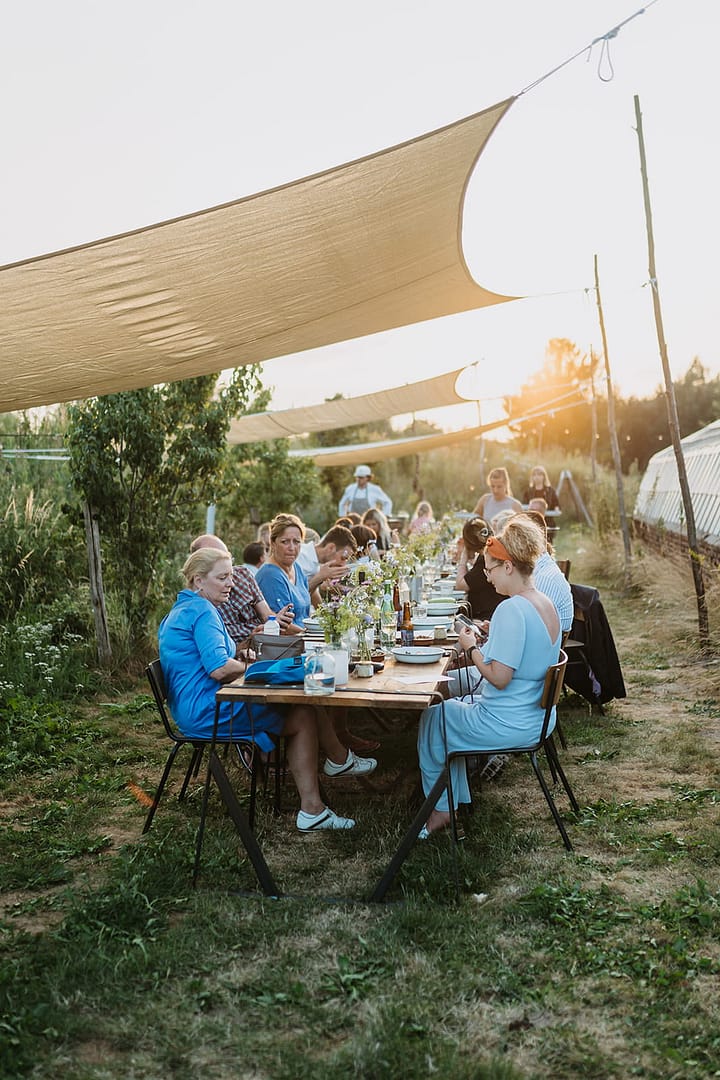 Gäste sitzen an einer langen Tafel unter Sonnensegeln und genießen ein gemeinsames Outdoor-Dinner in natürlicher Umgebung