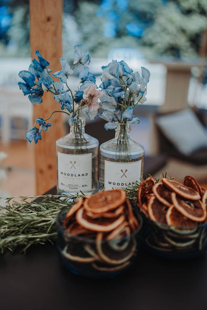 Detailaufnahme einer Bar mit Drinks, getrocknete Orangenscheiben und Blumenarrangement in Gin Flaschen bei einer Outdoor-Hochzeit, umgesetzt im Rahmen professioneller Eventplanung.