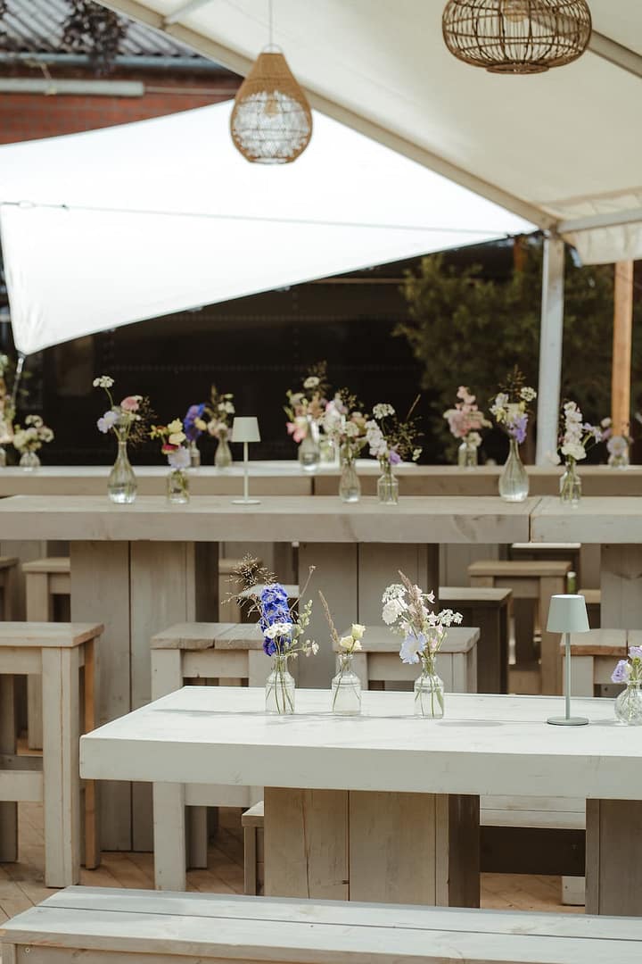 Minimalistisch und modern gedeckte Tafel mit feinen Glasvasen und Sommerblumen unter einer Zeltplane – Teil einer hochwertig geplanten Outdoor- und Zelthochzeit.