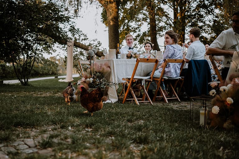 Gäste beim Hochzeitsdinner im Garten eines Ferienhauses mit Tieren wie Hund und Huhn, eingebettet in eine entspannte Wedding-Weekend-Atmosphäre.
