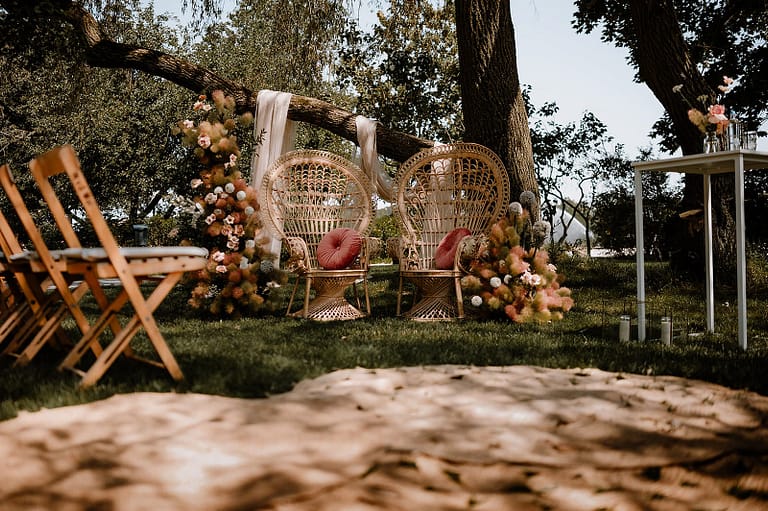 Zeremonie-Setup mit Peacock Chairs, floraler moderner Dekoration und Stoffbahnen für eine exklusive Gartenhochzeit an einem privaten Ferienhaus.