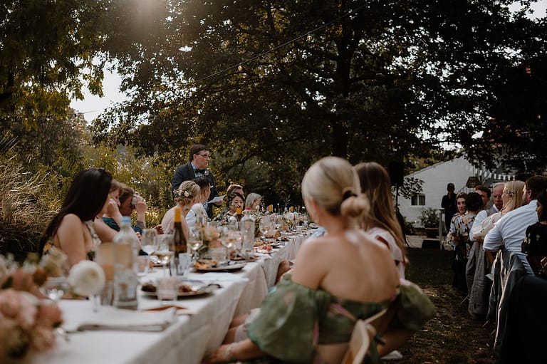 Gemeinsames Hochzeitsdinner mit Reden unter Bäumen und Lichterketten bei einer Outdoor-Hochzeit an einem privaten Ferienhaus.