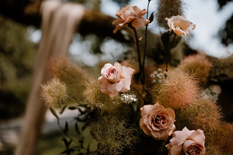 Moderne Hochzeitsdekoration mit roséfarbenen Rosen und wilden Gräsern als florales Detail einer hochwertig geplanten Outdoor- und Zelthochzeit an einem Ferienhaus.