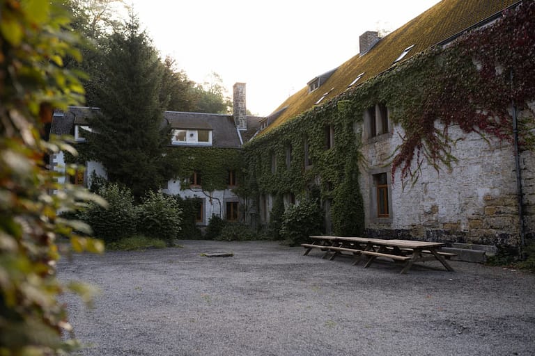 Ruhiger Innenhof einer naturnahen Hochzeitslocation mit Efeu bewachsenen Steinwänden und langen Holztischen – Vorbereitung für ein entspanntes Wedding Weekend.