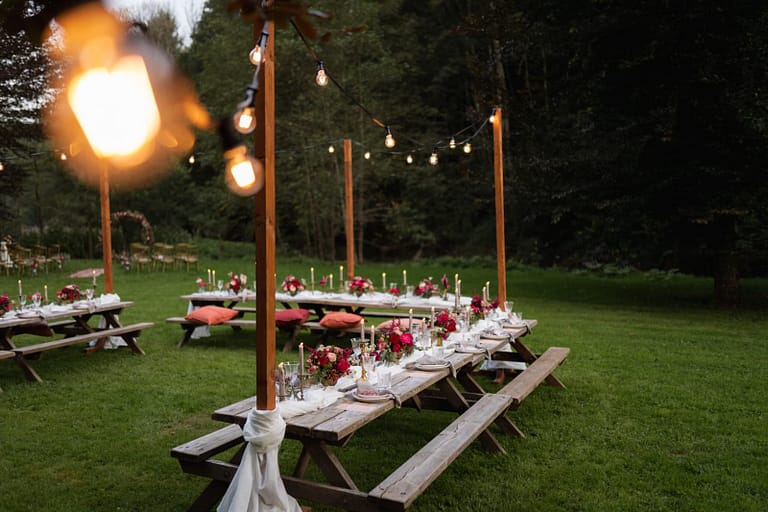 Kerzen, Blumen und gedeckte Holztafeln als Detail eines liebevoll geplanten Outdoor-Hochzeitsdinners mit Picknick Charakter.