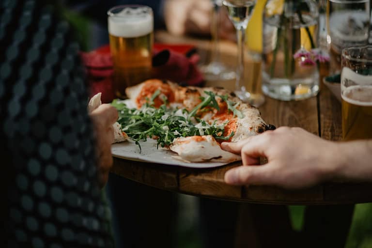 Gäste teilen Pizza und Getränke an Holztischen während einer ungezwungenen Hochzeitsfeier im Freien.