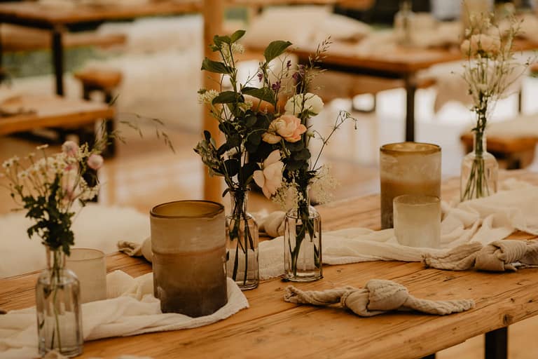 Detailaufnahme von hygge Blumenarrangements, natürlichen Textilien und Kerzen auf Holztischen - den Scandi Look abbildend - bei einer entspannten Hochzeit im Freien.