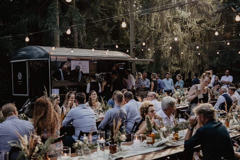 Geselliges Outdoor-Dinner bei einer Hochzeit im Wald mit Foodtruck, langen Tafeln und stimmungsvoller Beleuchtung – Teil eines entspannten Wedding Weekends in natürlicher Off-Location.