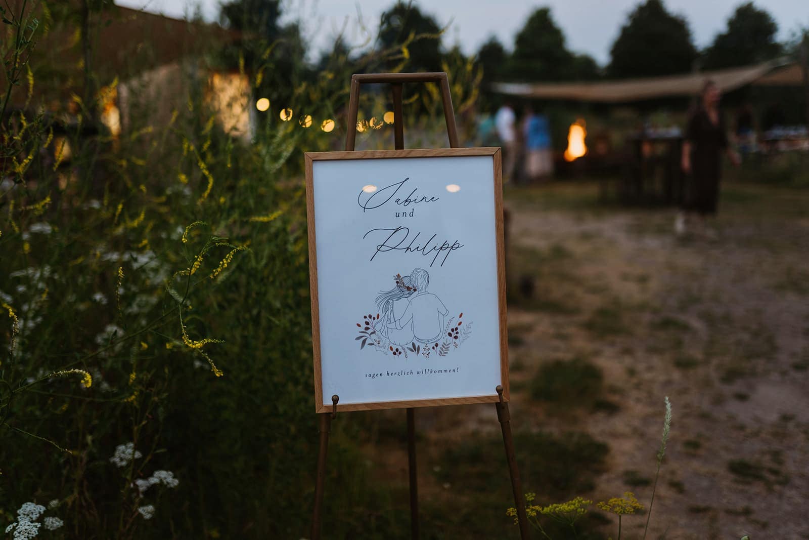 Willkommensschild bei einer Outdoor Hochzeit in der Abendstimmung mit Lichterketten und Gästen im Hintergrund